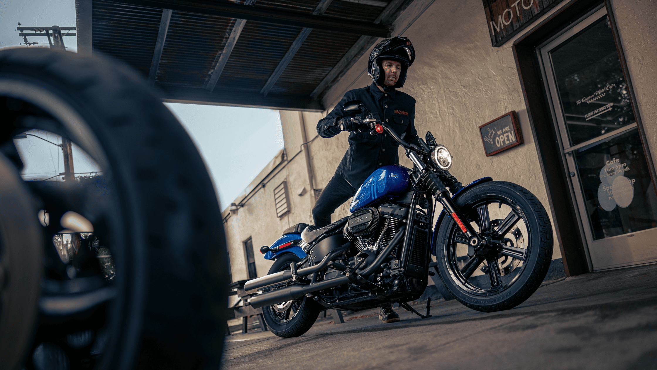 Rider standing beside the 2024 Harley-Davidson® Street Bob® 114 motorcycle in vibrant blue outside a shop in Ormond Beach, FL, capturing the bike's rugged design and urban appeal.